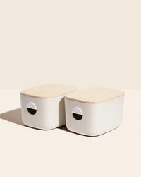 Two Cream Medium Storage Bins with wooden lids on a cream background. 
