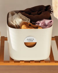 The Open Spaces Bin Label on a Cream Medium Storage Bin with cloths on a wooden rack.