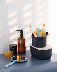 Small Navy Storages Bins with beauty products and one with a wooden lid on top of each other on a bathroom shelf.