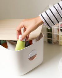 A hand taking out a bottle from the Cream Medium Storage bin with half open wooden lid.