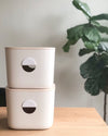 Two Cream Medium Storage Bins with wooden lids stacked up on top of each other. 