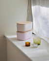 Two Small Light Pink Storages Bins with wooden lids stacked on top of each other next to candles on a white countertop..