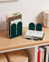 Two Open Spaces Dark Green Tortuga Bookends with books on a wooden shelf with other items on it. 
